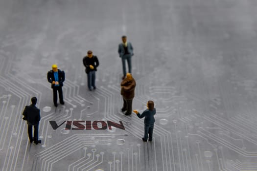 Miniature figurines posed as business people in a meeting over abstract futuristic circuit board with text Vision