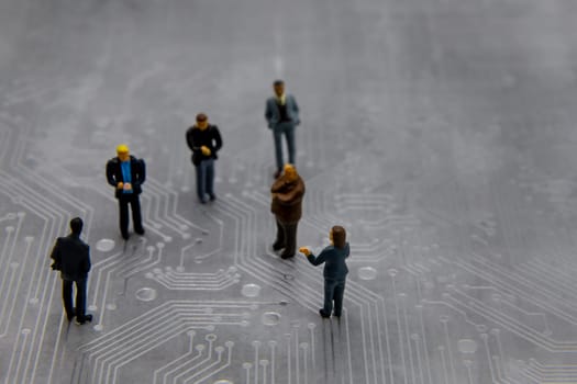 Miniature figurines posed as business people in a meeting over abstract futuristic circuit board, future business concept