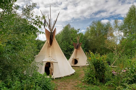 Two Indian tipis stand in a meadow between trees and bushes in the sunshine