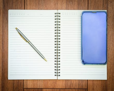 Top view image of open notebook with blank pages and cellphone on wooden table background for adding text or mockup
