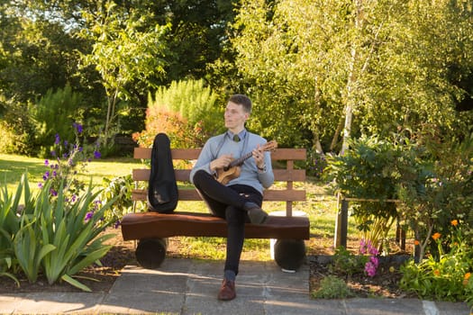 Young man sitting on bench in the park and playing ukulele.