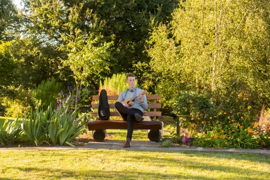 Young man sitting on bench in the park and playing ukulele.