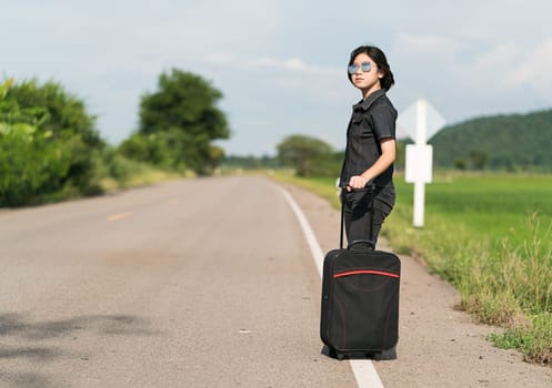 Young asian woman short hair and wearing sunglasses with luggage hitchhiking along a road in countryside Thailand