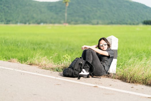 Young asian woman short hair and wearing sunglasses sit with backpack hitchhiking along a road wait for help in countryside Thailand