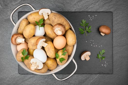 Raw potatoes, eggs, halved and whole brown mushrooms, coarse salt grains and parsley leaves in a white enamel sieve on a slate slap. Flat lay.