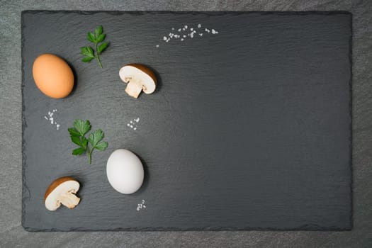 Mushroom Omelet ingredients with two half brown mushrooms, a brown and a white egg, two parsley leaves and coarse salt grains on a slate slab. With copy space.Flat lay.