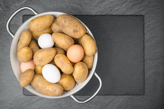 Raw potatoes and eggs in a white enamel sieve on a slate slap. Flat lay.