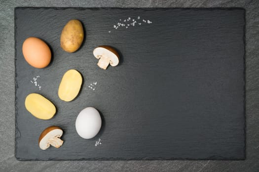 Mushroom Tortilla ingredients with two half brown mushrooms, a brown and a white egg, halved potatoes and coarse salt grains on a slate slab. With copy space. Flat lay.