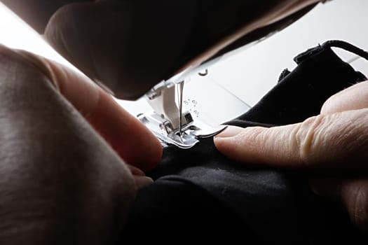 High contrast close up image of a black face covering or mask being fed into a sewing machine by older aged hands.