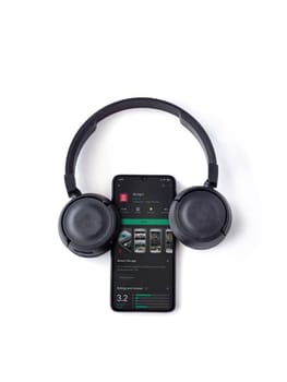 Lod, Israel - July 8, 2020: Black mobile smartphone with Reshet 13 app play store page and wireless headphones on a white background. Top view flat lay with copy space.