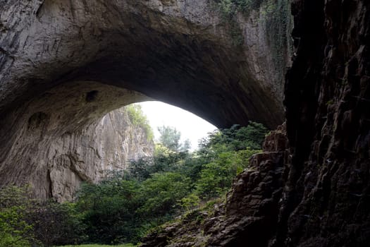 perspective view of the Devetaki cave in Bulgaria with green forest vegetation and round opening in the rock ceiling