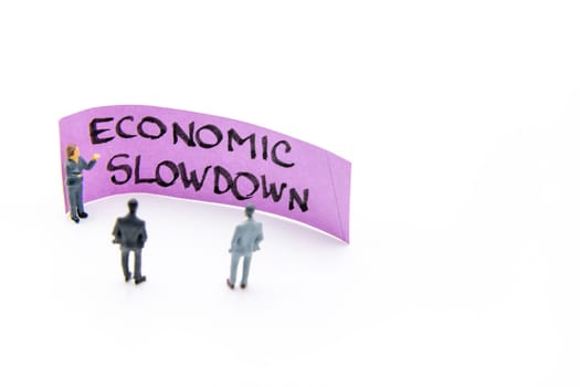 Presentation meeting with miniature figurines posed as business people standing in front of post-it note with Economic Slowdown handwritten message in background, copy space on the right