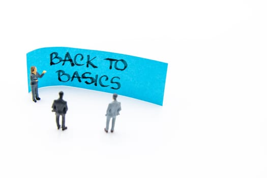 Presentation meeting with miniature figurines posed as business people standing in front of post-it note with Back To Basics handwritten message in background, copy space on the right