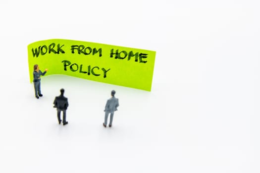 Presentation meeting with miniature figurines posed as business people standing in front of post-it note with Work From Home Policy handwritten message in background, copy space on the right