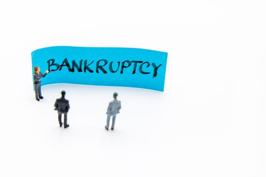 Presentation meeting with miniature figurines posed as business people standing in front of post-it note with Bankruptcy handwritten message in background, copy space on the right
