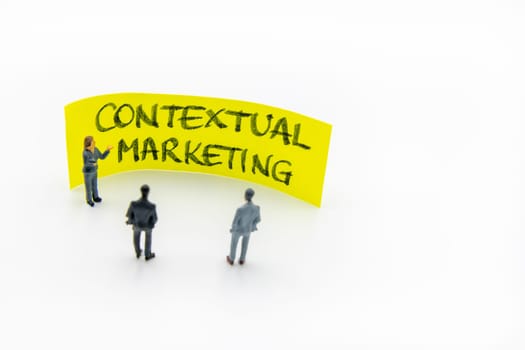 Presentation meeting with miniature figurines posed as business people standing in front of post-it note with Contextual Marketing handwritten message in background, copy space on the right