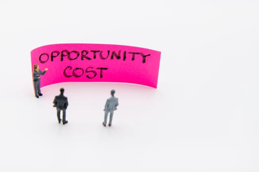 Presentation meeting with miniature figurines posed as business people standing in front of post-it note with Opportunity Cost handwritten message in background, copy space on the right