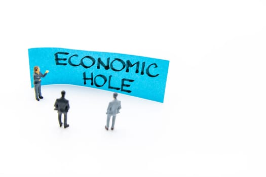 Presentation meeting with miniature figurines posed as business people standing in front of post-it note with Economic Hole handwritten message in background, copy space on the right