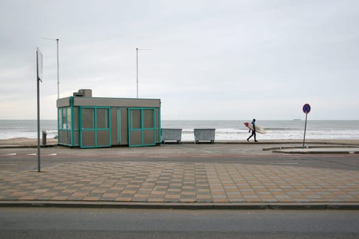 Lone surfer in neoprene swimsuit walking on pavement to empty beach on a cold winter overcast day. The Hague, Netherlands