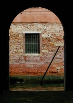 Dark interior opens through archway to ancient red brick wall and window with bars. Beautiful green and red colors and texture.
