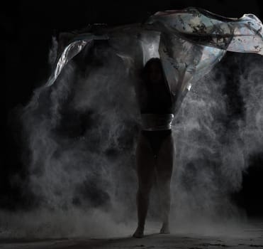 young athletic girl with long hair dances with a piece of cloth in a cloud of white flour, low key
