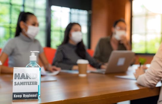 Signage of Hand sanitizer alcohol gel in meeting room with background of Asian team business people working and brainstorm. They wear face mask in new normal office to prevent covid-19 virus spreading