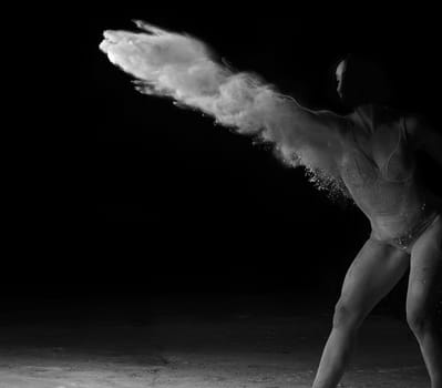 beautiful caucasian woman in a black bodysuit with a sports figure is dancing in a white cloud of flour on a black background, black white toning