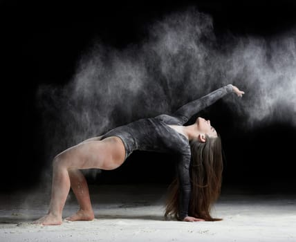 young beautiful caucasian woman with long hair is dressed in a sports black bodysuit stands in a bent pose and throws white flour, black background, gymnastic exercises