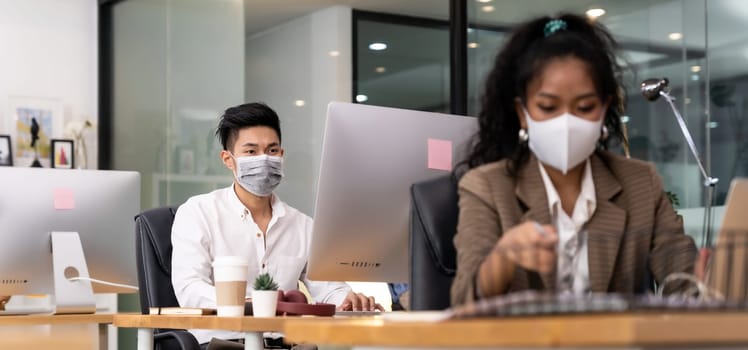 Panorama asian Office employee with protective face mask working in new normal office with social distance practice to american african colleague in foreground prevent coronavirus COVID-19 spreading.