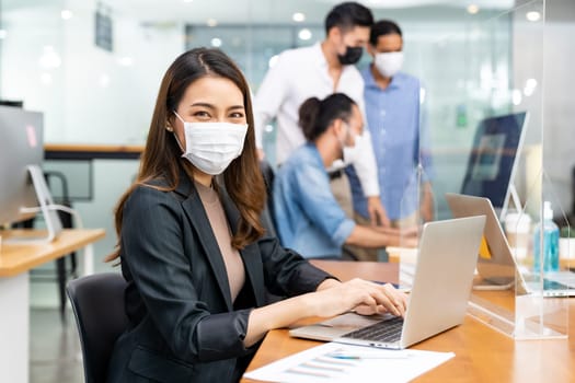 Portrait of asian office employee businesswoman wear protective face mask work in new normal office with interracial team in background as social distance practice prevent coronavirus COVID-19.