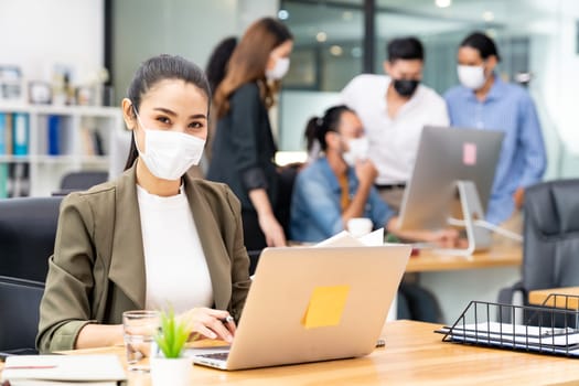 Portrait of asian office employee businesswoman wear protective face mask work in new normal office with interracial team in background as social distance practice prevent coronavirus COVID-19.
