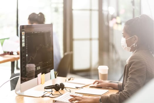 Mixed race of African black and asian businesswoman office worker wear protective face mask working in new normal office with social distance between table to prevent coronavirus COVID-19 spreading.