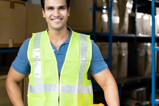Portrait of Indian asian warehouse worker with safety vest stand and hold yellow helmet in warehouse distribution center environment. Using in business warehouse and logistic concept.
