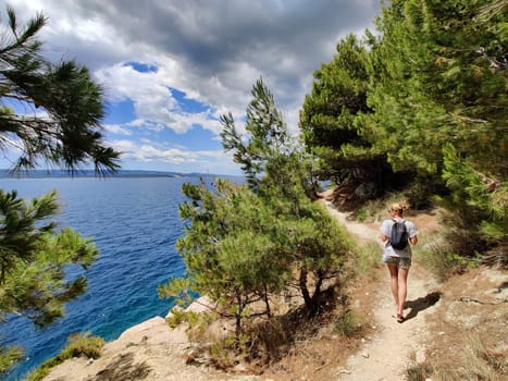 Young active feamle tourist wearing small backpack walking on coastal path among pine trees looking for remote cove to swim alone in peace on seaside in Croatia. Travel and adventure concept.