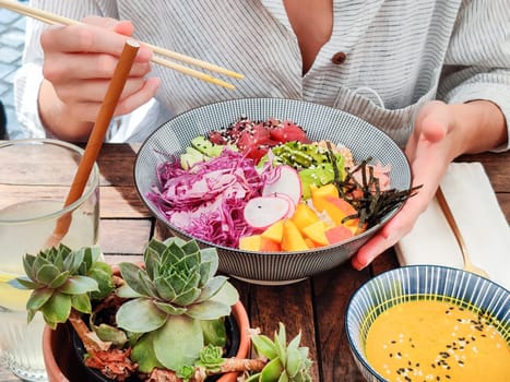 Woman eating tasty colorful healthy natural organic vegetarian Hawaiian poke bowl using asian chopsticks on rustic wooden table. Healthy natural organic eating concept.