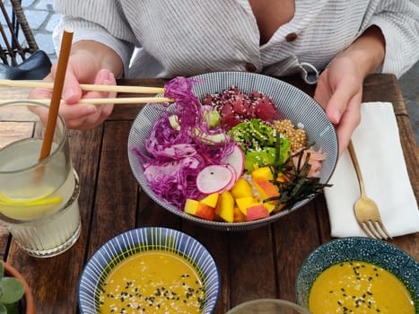 Woman eating tasty colorful healthy natural organic vegetarian Hawaiian poke bowl using asian chopsticks on rustic wooden table. Healthy natural organic eating concept.