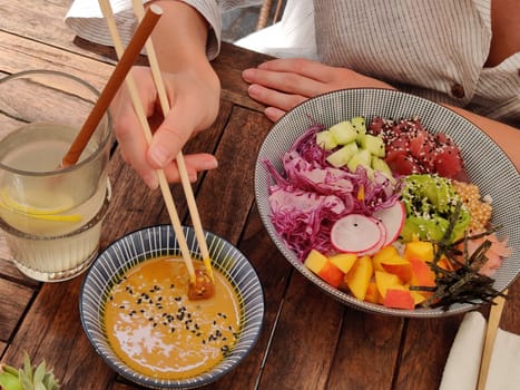 Woman eating tasty colorful healthy natural organic vegetarian Hawaiian poke bowl using asian chopsticks on rustic wooden table. Healthy natural organic eating concept.