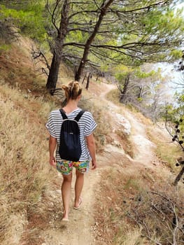 Young active feamle tourist wearing small backpack walking on coastal path among pine trees looking for remote cove to swim alone in peace on seaside in Croatia. Travel and adventure concept.