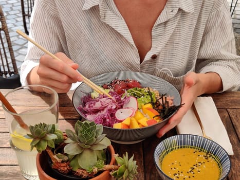 Woman eating tasty colorful healthy natural organic vegetarian Hawaiian poke bowl using asian chopsticks on rustic wooden table. Healthy natural organic eating concept.