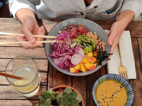 Woman eating tasty colorful healthy natural organic vegetarian Hawaiian poke bowl using asian chopsticks on rustic wooden table. Healthy natural organic eating concept.