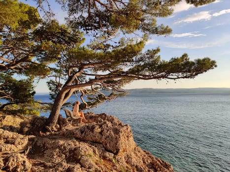 Pensive woman on vacations, sitting and relaxing under large pine tree on bench by dip blue sea enjoying beautiful sunset light in Brela, Makarska region, Dalmatia, Croatia.