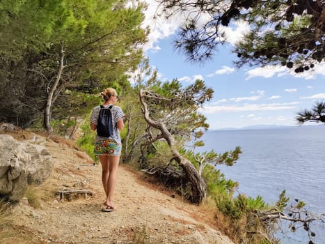 Young active feamle tourist wearing small backpack walking on coastal path among pine trees looking for remote cove to swim alone in peace on seaside in Croatia. Travel and adventure concept.