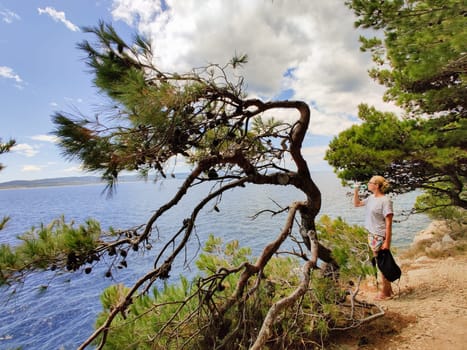Young active feamle tourist taking a break, drinking water, wearing small backpack while walking on coastal path among pine trees looking for remote cove to swim alone in peace on seaside in Croatia.