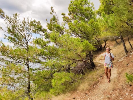 Young active feamle tourist wearing small backpack walking on coastal path among pine trees looking for remote cove to swim alone in peace on seaside in Croatia. Travel and adventure concept.