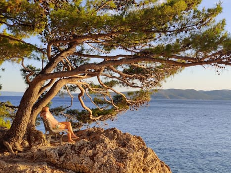 Pensive woman on vacations, sitting and relaxing under large pine tree on bench by dip blue sea enjoying beautiful sunset light in Brela, Makarska region, Dalmatia, Croatia.