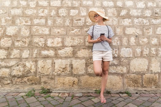 Beautiful young female tourist woman standing in front of old textured stone wall at old Mediterranean town, smiling, holding, smart phone to network on vacationes. Copy space.