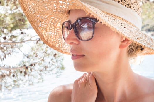 Close up portrait of no makeup natural beautiful woman wearing straw sun hat and sun glasses on the beach in shade of a pine tree.