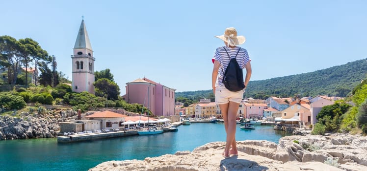 Woman traveler wearing straw summer hat and backpack, standing at edge of the rocky cliff enjoying beautiful panoramic view of Veli Losinj, Losinj island, Croatia.