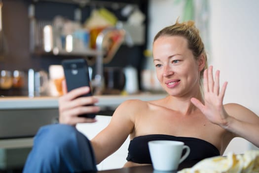 Young smiling cheerful woman indoors at home kitchen using social media on phone for video chatting and staying connected with her loved ones. Stay at home, social distancing lifestyle.