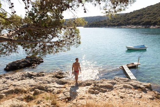 Young sporty man wearing red speedos tanning and realaxing on wild cove of Adriatic sea on a beach in shade of pine tree.